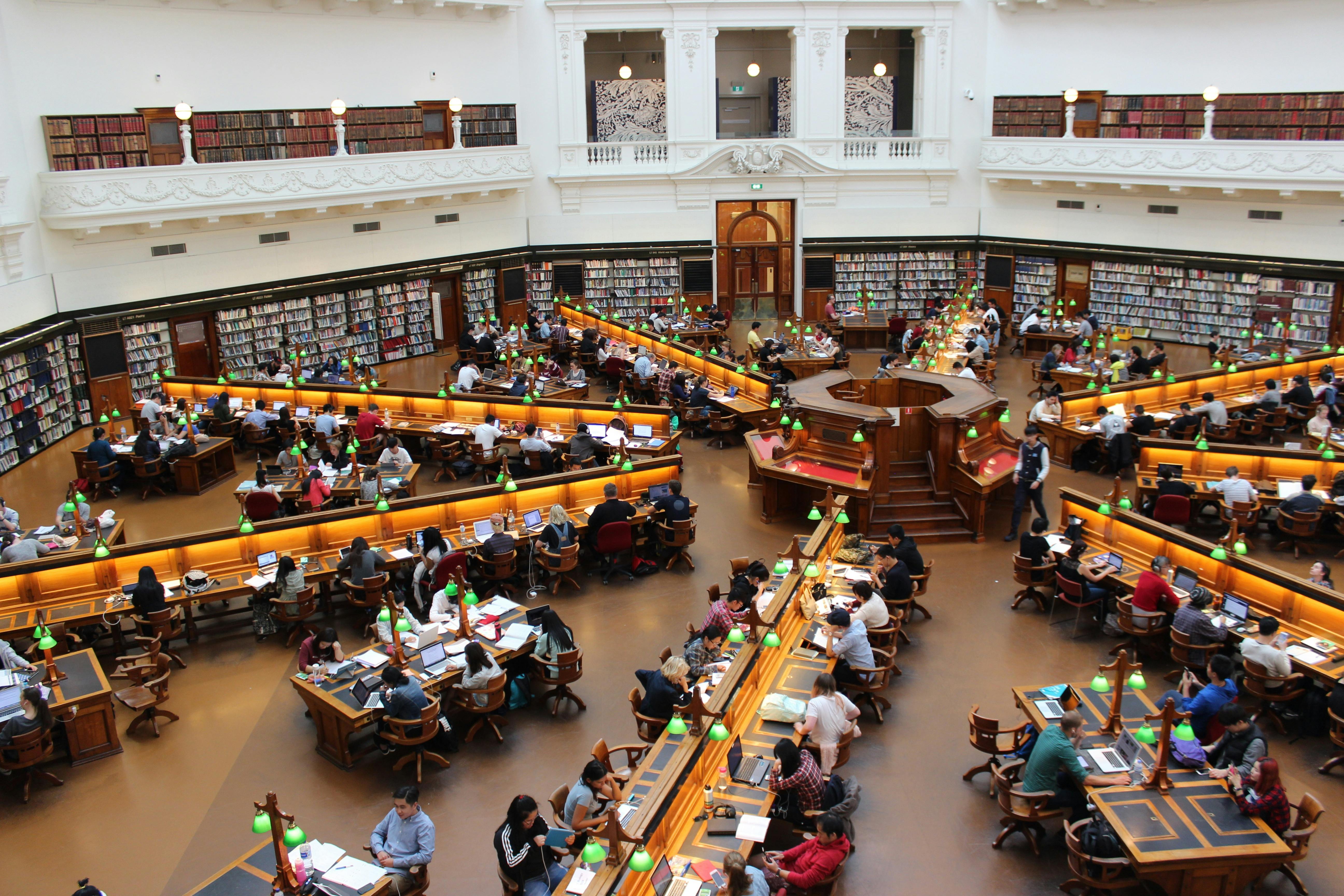 Large library reading room with rows of wooden desks, green desk lamps, and people studying or working on laptops.