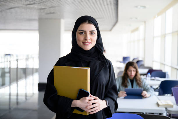 Smiling young woman in a black hijab holding a yellow folder and smartphone in a modern study area.