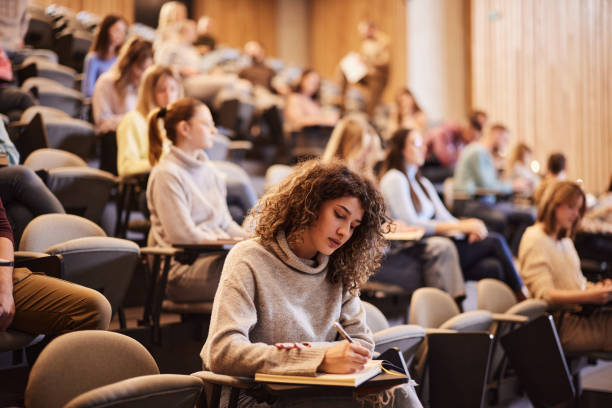 Young woman with curly hair writing notes in a large lecture hall filled with students.