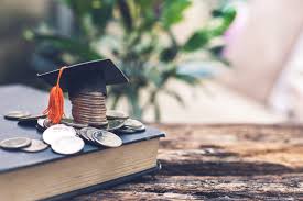 Stack of coins wearing a graduation cap placed on a closed book with scattered coins around it on a wooden surface.