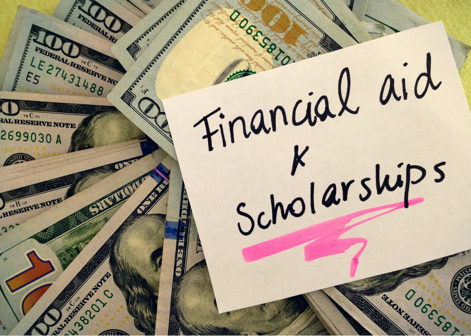 Handwritten note reading 'Financial aid & Scholarships' on pink-highlighted paper laid over spread of US hundred-dollar bills.