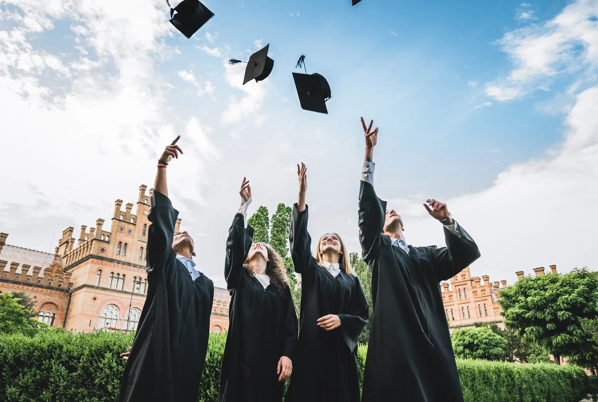 Four graduates in black caps and gowns throwing their mortarboards into the air outdoors with historic buildings and trees in the background.