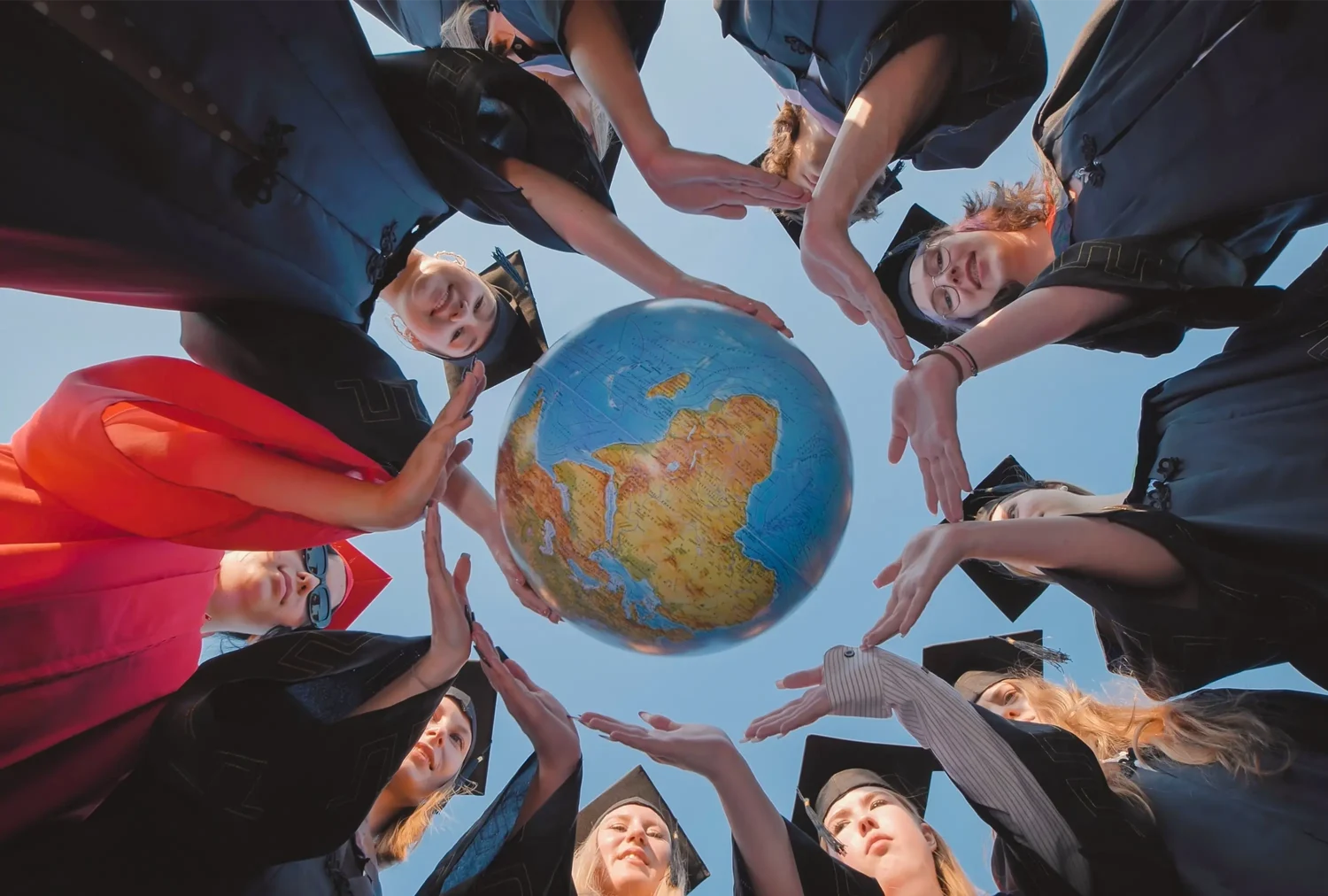 Graduates in caps and gowns standing in a circle outdoors, holding a globe above them against a clear sky.