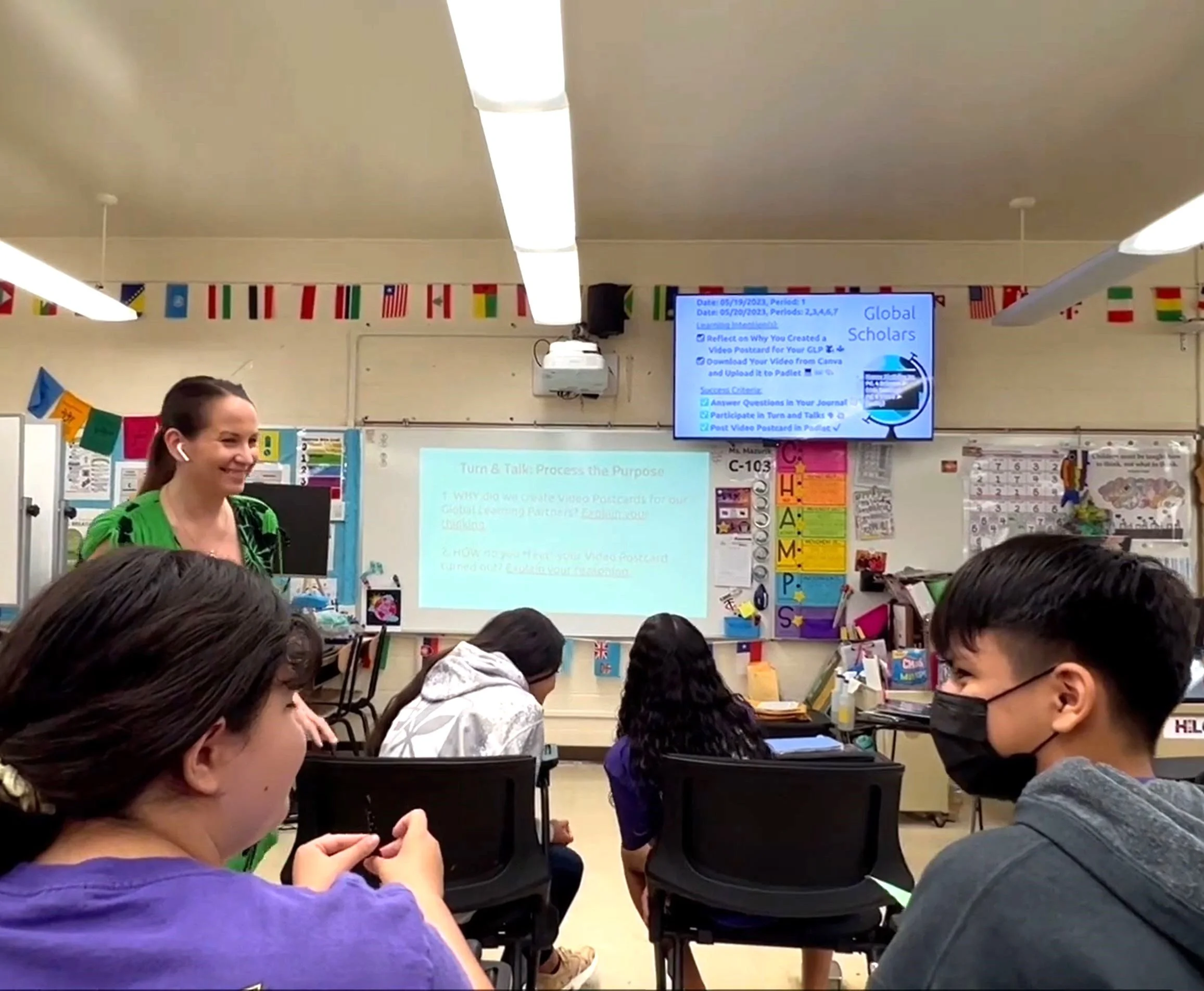 Classroom scene with a smiling teacher standing and students sitting on chairs facing a projector screen and a monitor displaying instructions titled Global Scholars.