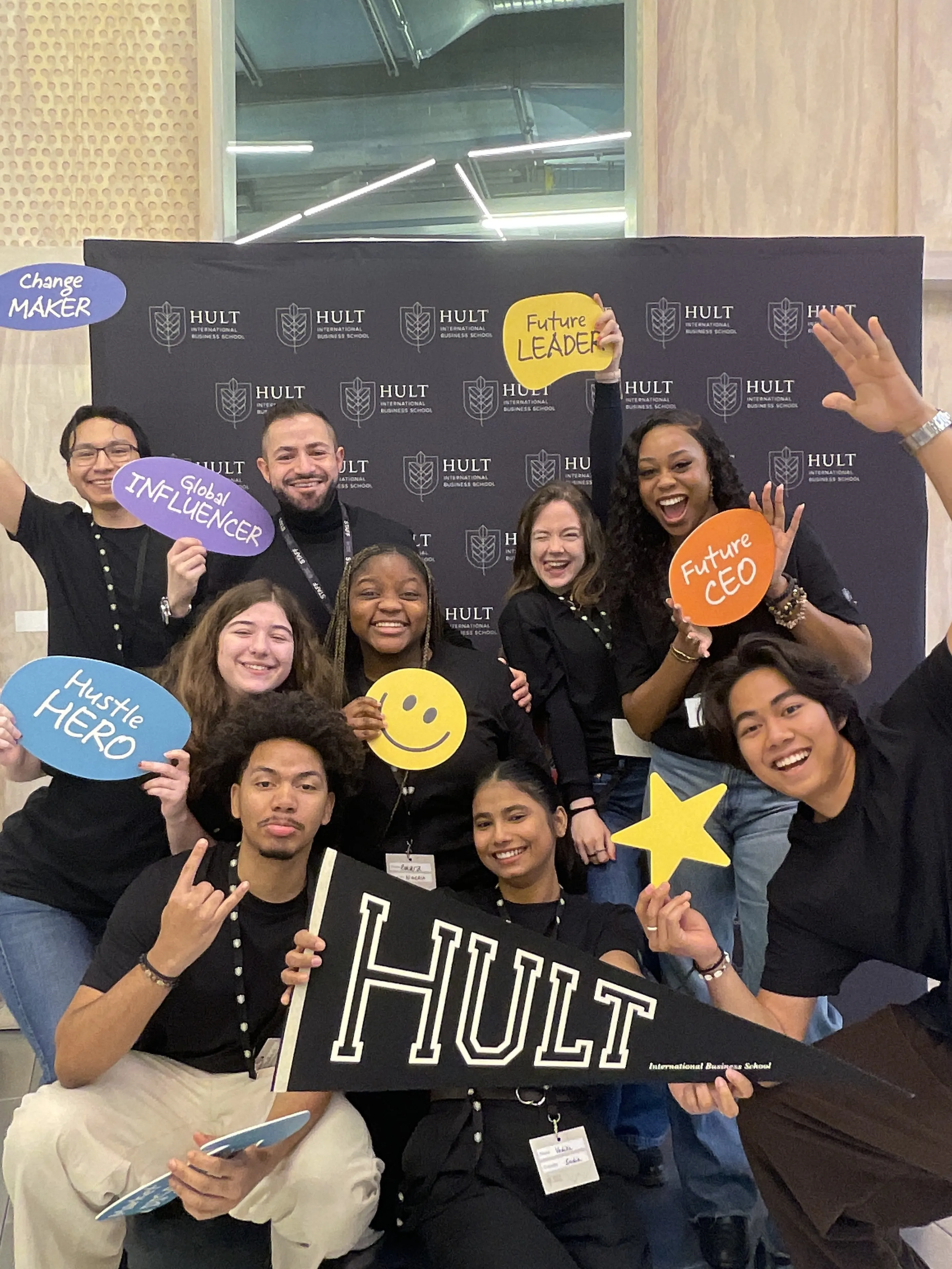 Group of smiling diverse young adults holding signs with motivational titles like Future Leader, Future CEO, Hustle Hero, and Global Influencer in front of a Hult International Business School backdrop.