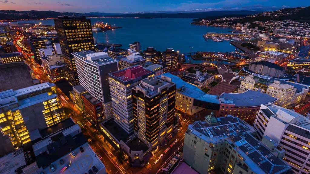 Aerial view of a city at dusk with illuminated buildings and streets next to a large body of water.