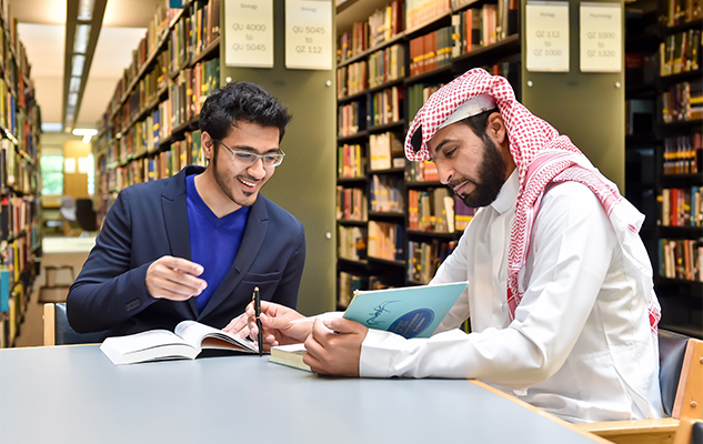 Two men studying together at a library table surrounded by bookshelves, one wearing a suit and glasses and the other in traditional Middle Eastern attire.