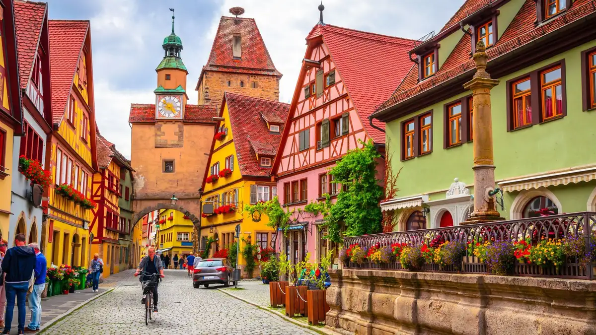 Cobbled street lined with colorful half-timbered houses and flower boxes, featuring a clock tower and people walking and cycling.