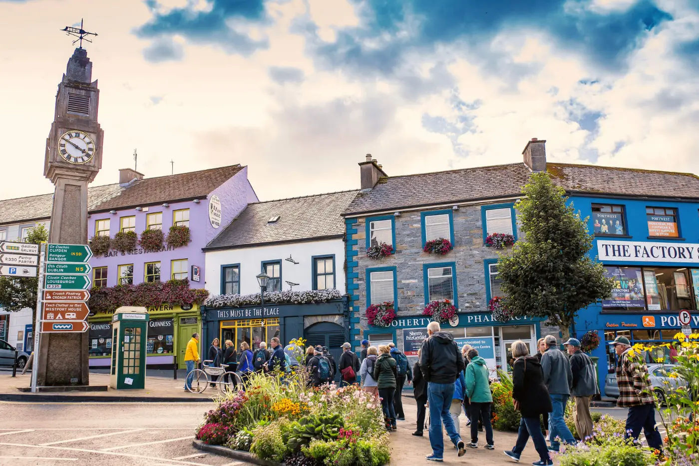 People walking along a street with colorful buildings, a clock tower, and a garden in a quaint town.