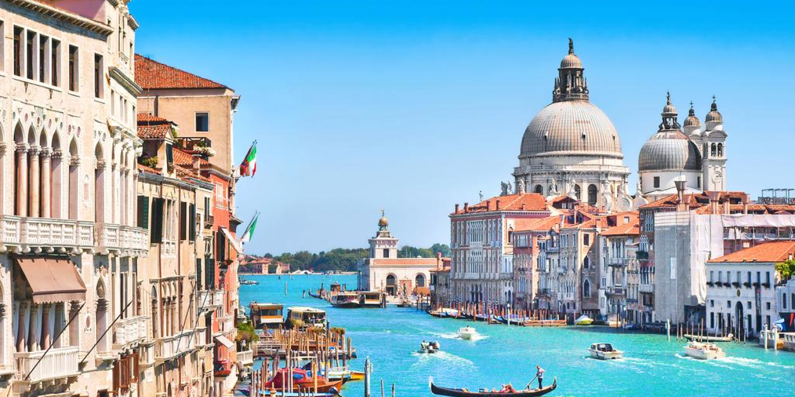 Vibrant canal in Venice with gondolas and boats, historic buildings lining the water, and the Santa Maria della Salute church under a clear blue sky.