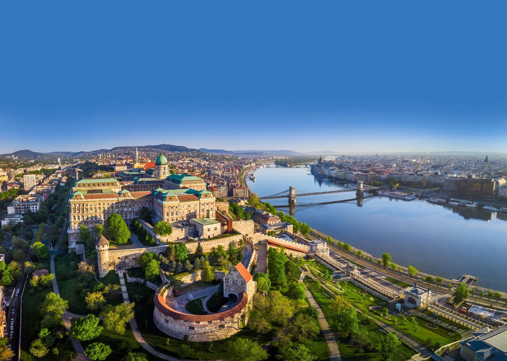 Aerial view of Buda Castle and the Chain Bridge over the Danube River in Budapest, Hungary, with cityscape and hills in the background under a clear blue sky.