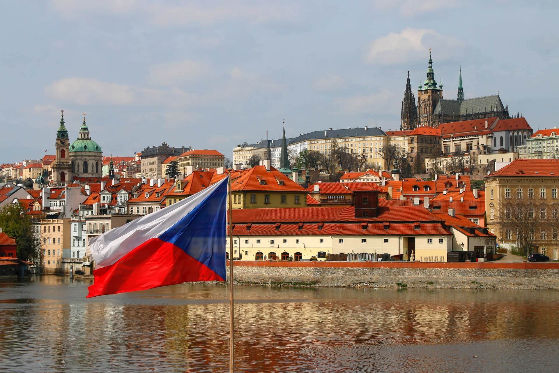 Czech Republic flag waving over river with historic buildings and Prague Castle in the background under a partly cloudy sky.