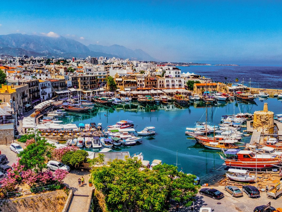 A vibrant harbor with boats docked in calm turquoise water, surrounded by buildings, mountains in the background, and a clear blue sky.