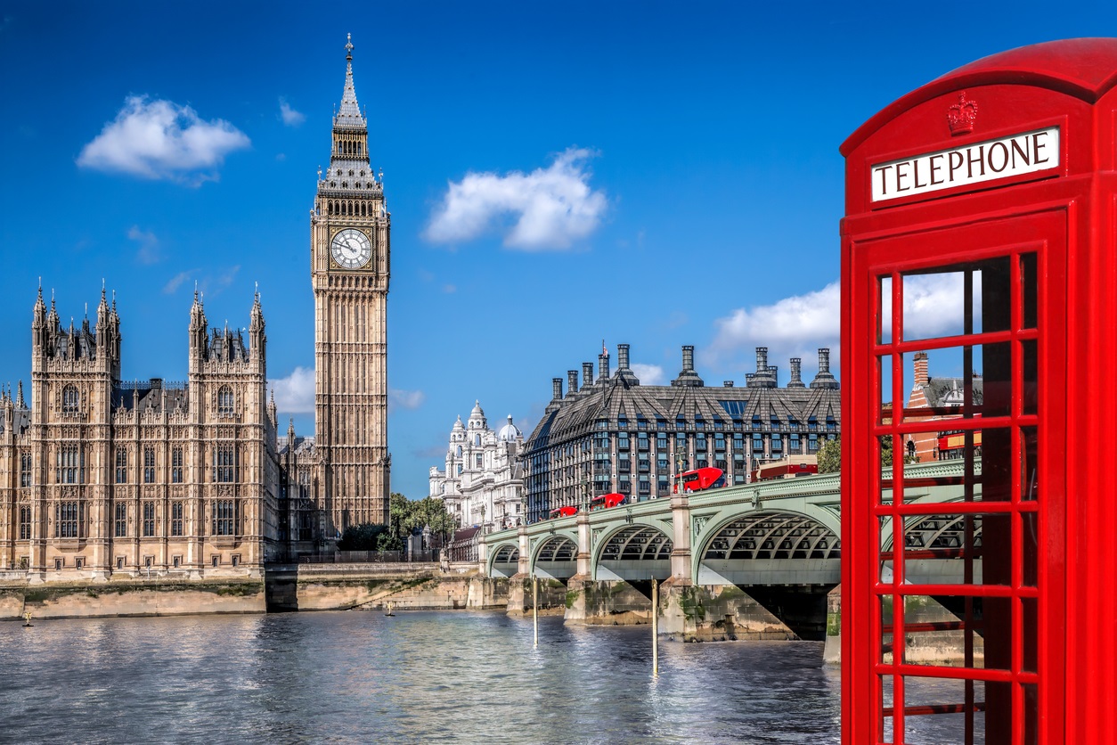 Red British telephone booth with Big Ben, the Houses of Parliament, and Westminster Bridge in London under a blue sky.