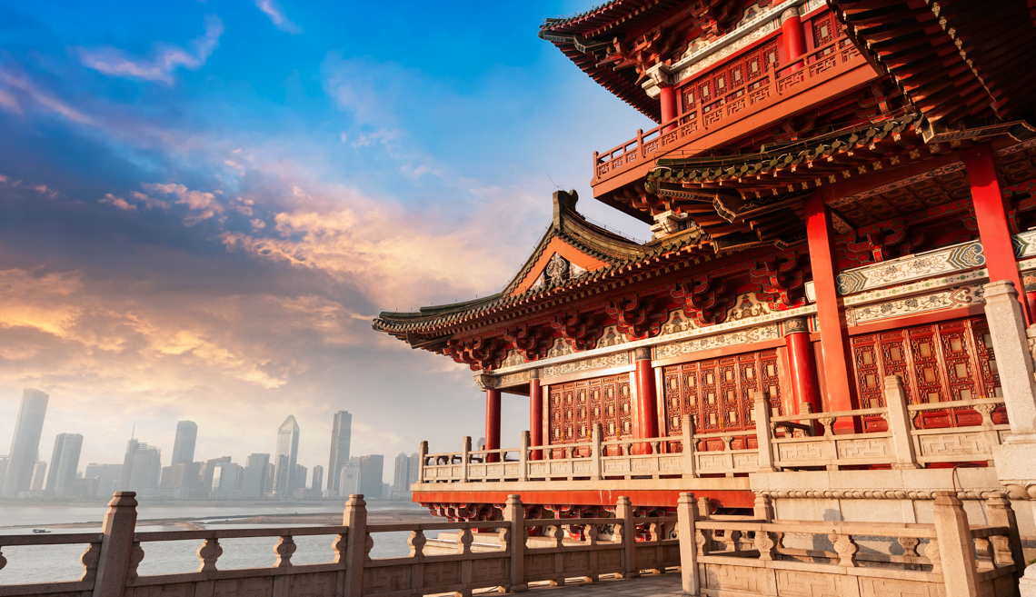 Traditional red Chinese temple with ornate roof overlooking a river and modern city skyline under a colorful sunset sky.