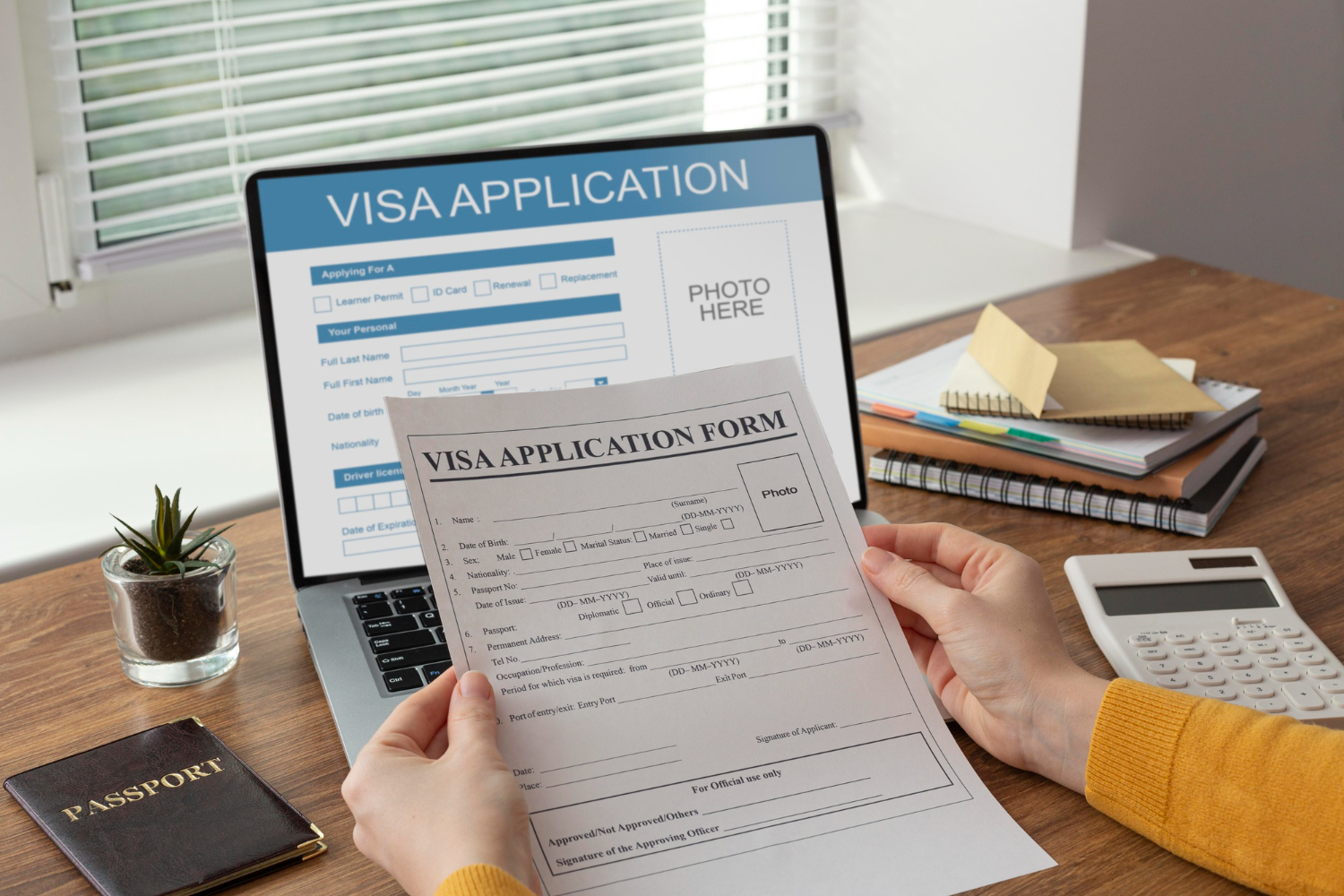 Person holding a visa application form paper with a laptop displaying an online visa application form, a passport, a calculator, and notebooks on a wooden desk.