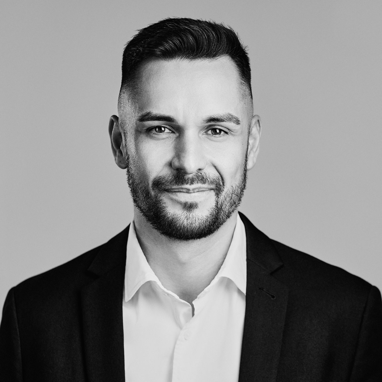Black and white headshot of Guenther Petelin with short dark hair, beard, white shirt, and dark blazer smiling slightly.