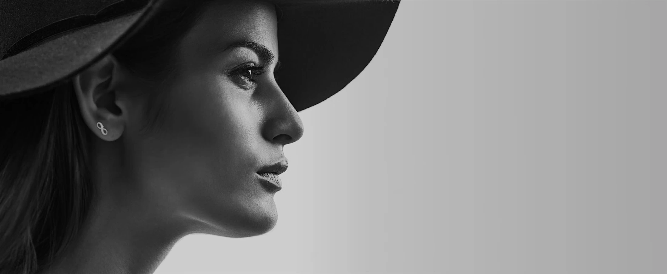 Black and white side profile of a woman wearing a wide-brimmed hat and infinity symbol earrings.