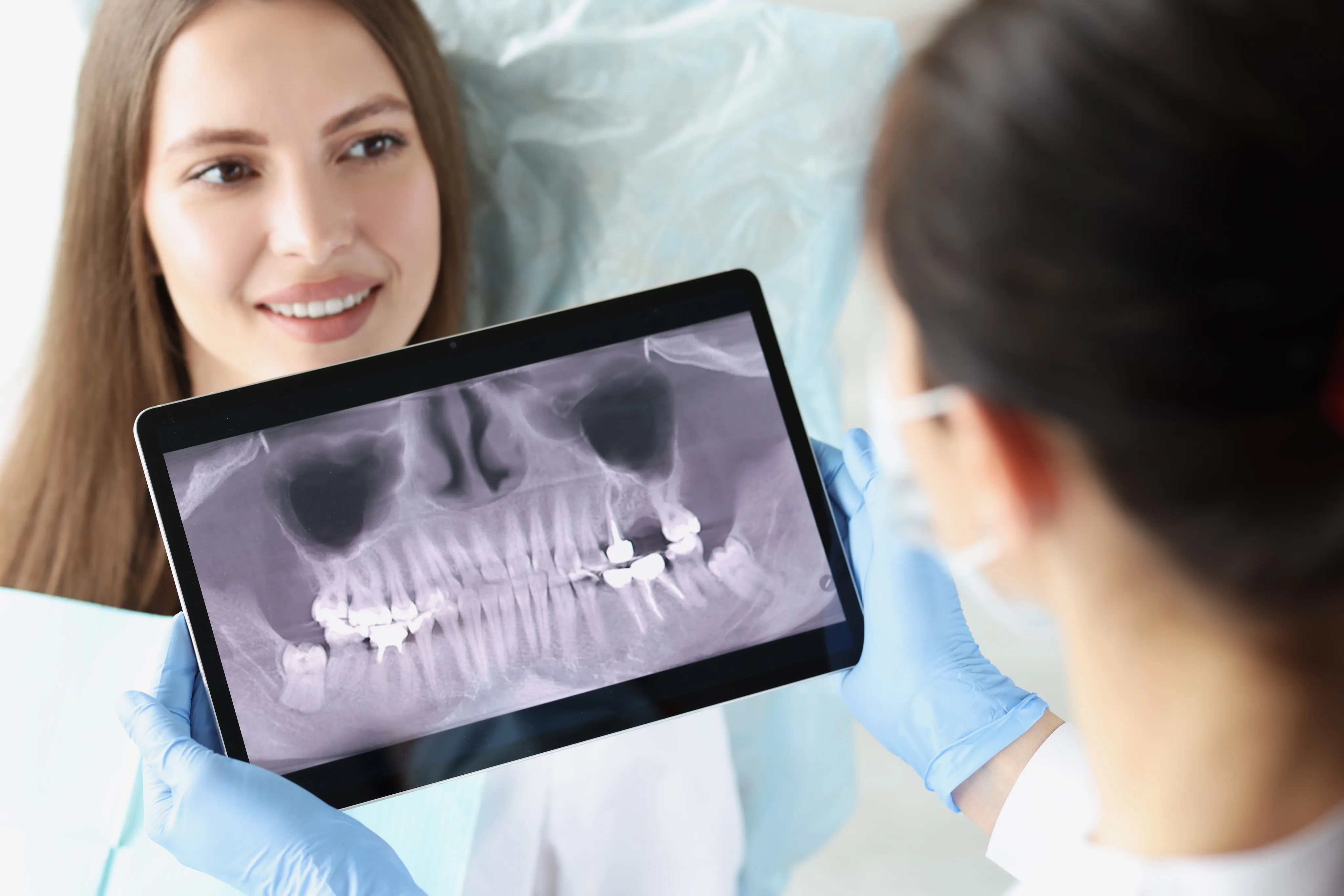 Dentist wearing blue gloves shows a dental X-ray on a tablet to a smiling female patient.
