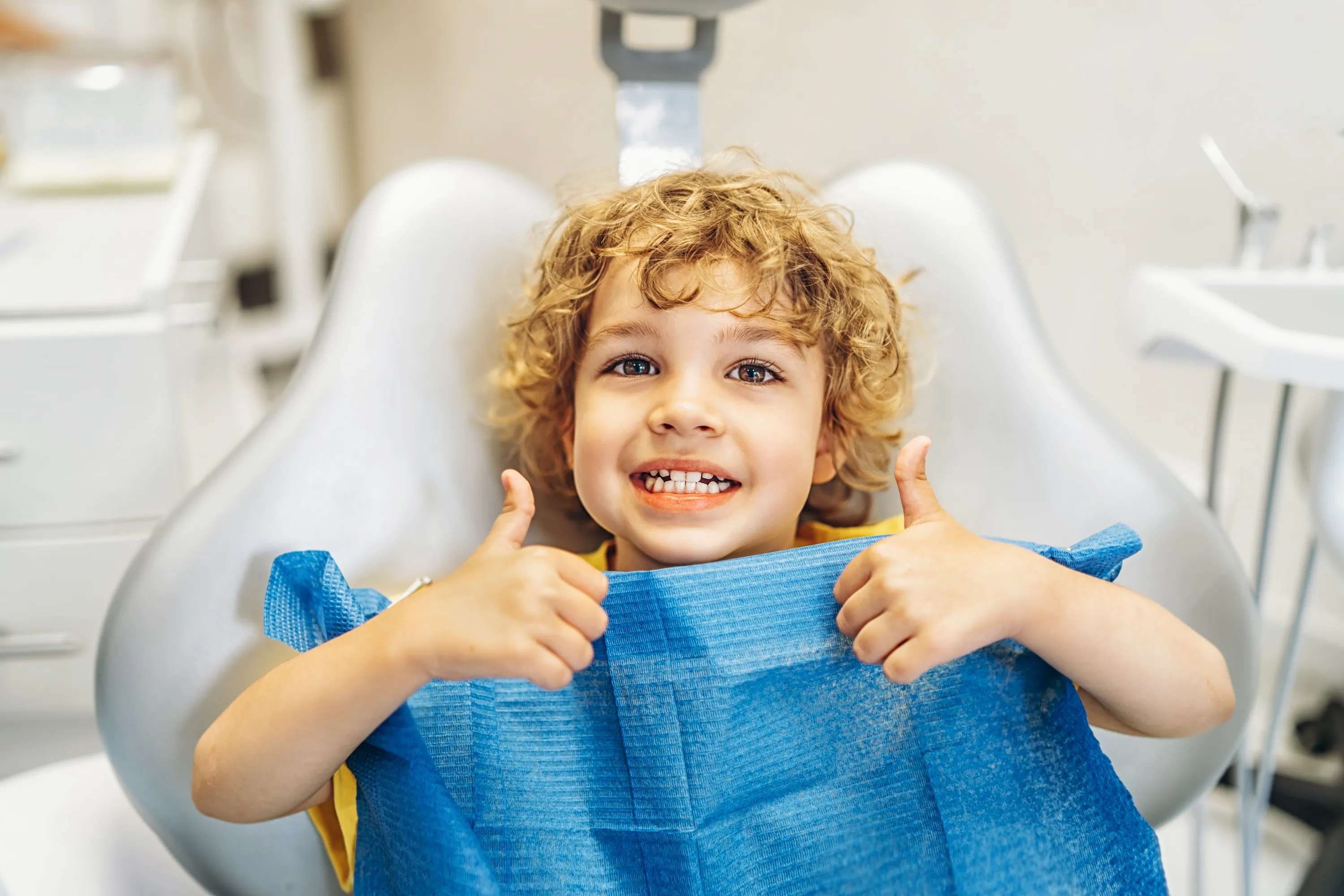 Smiling young child with curly hair sitting in a dental chair giving thumbs up with both hands.