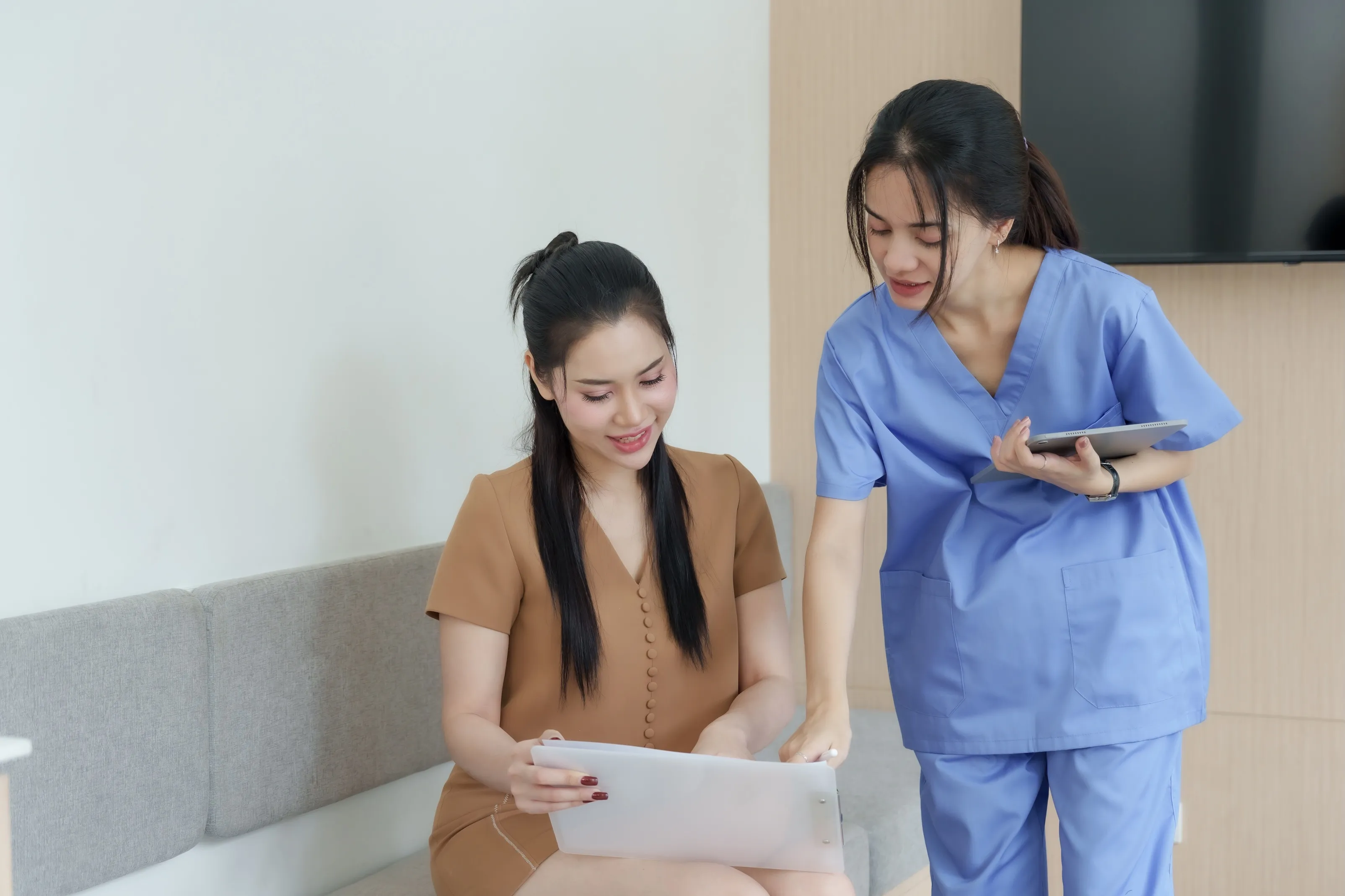 Healthcare professional in blue scrubs discussing a document with a seated woman in a brown dress.