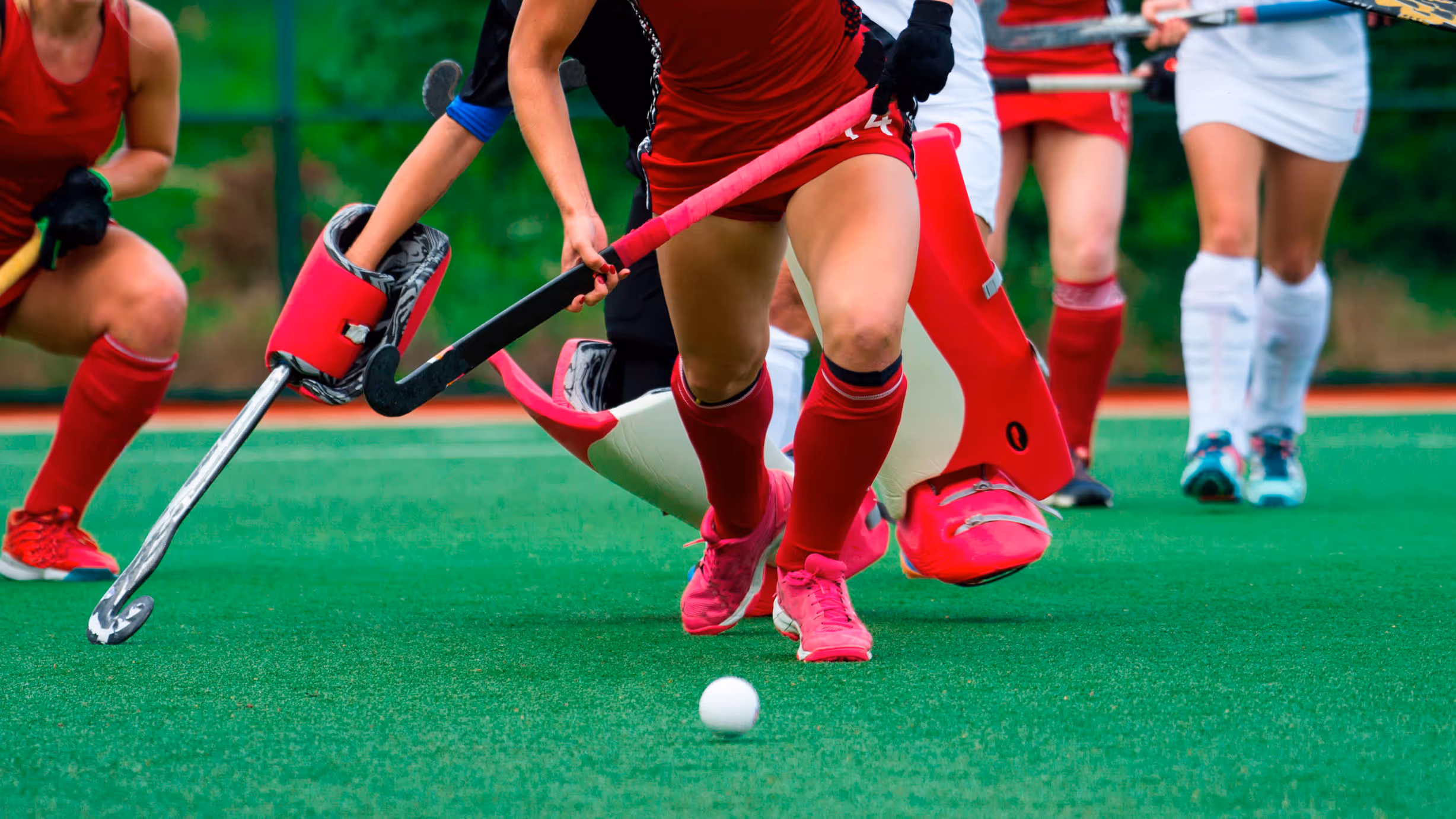 Field hockey players in red and white uniforms compete for the ball on a green field. The focus is on legs and sticks, conveying intensity and motion.