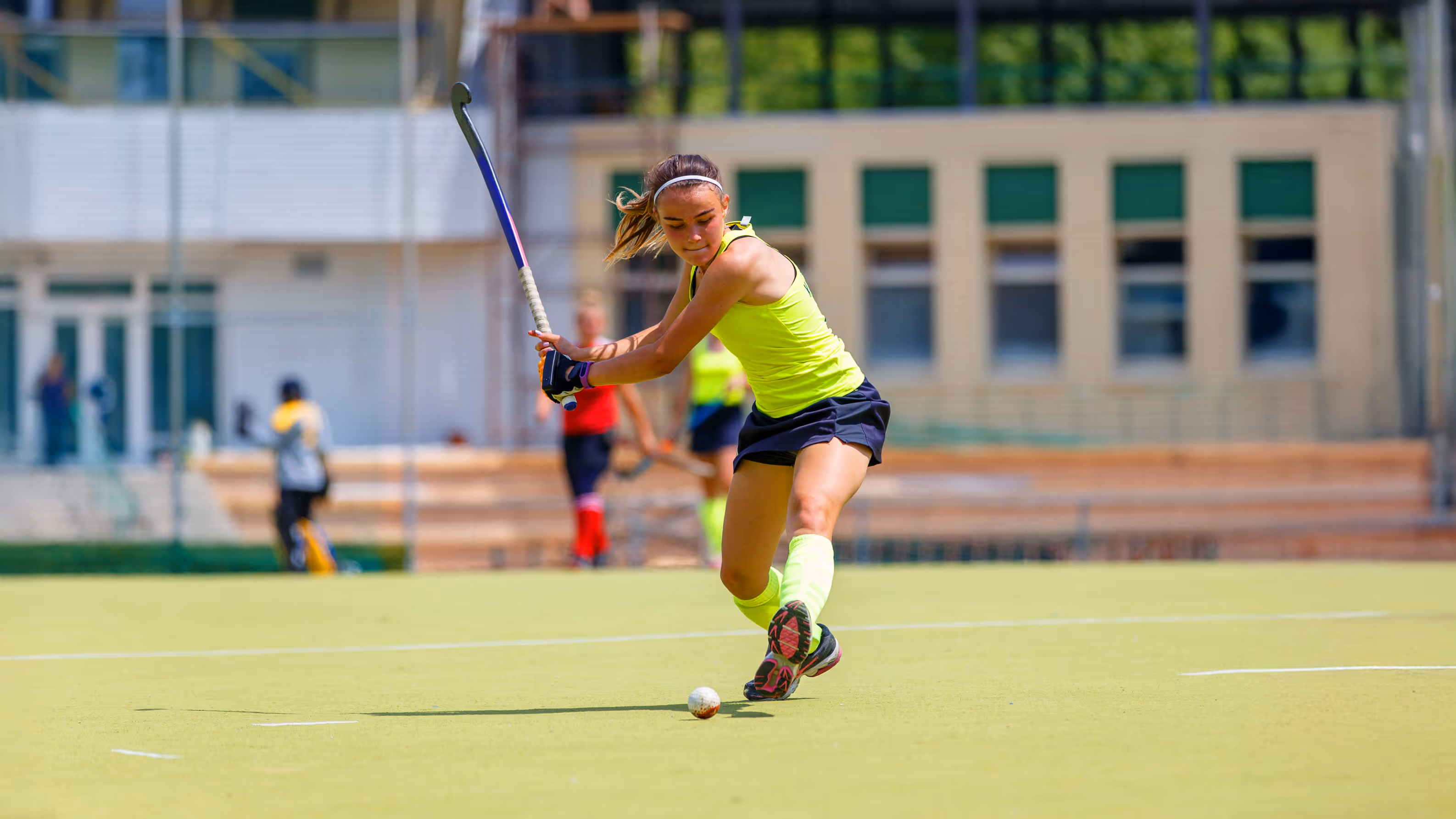 A female field hockey player in a neon yellow tank top concentrates on hitting a ball with her stick on a sunny day. The field and background are blurred.