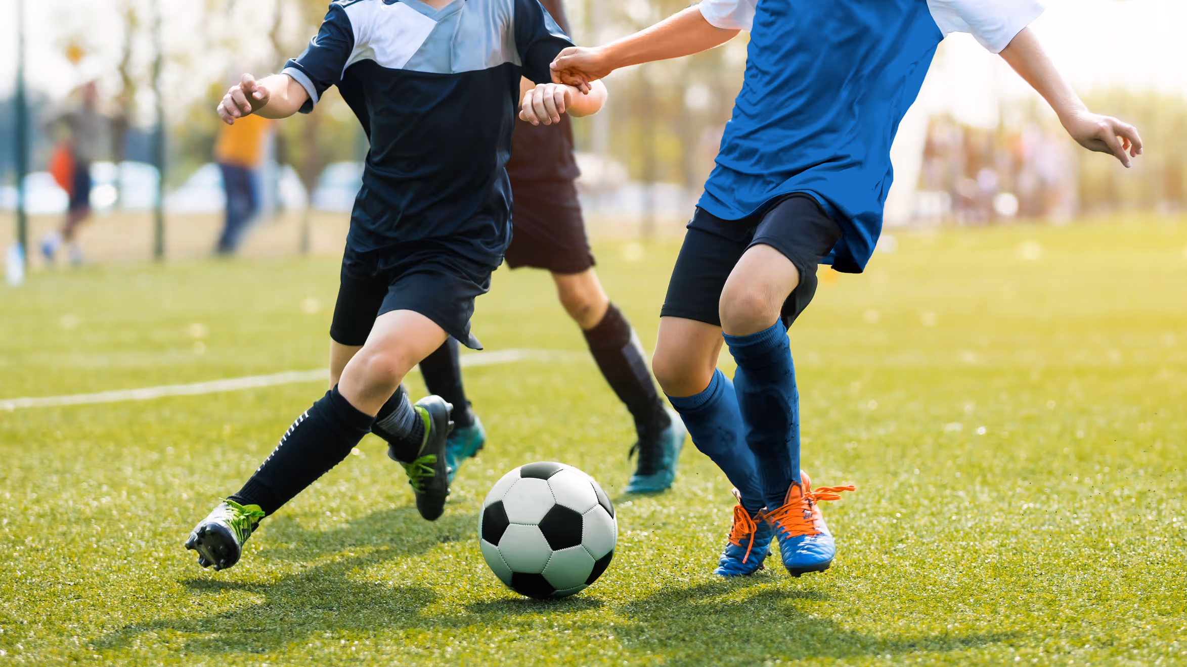 Two youth soccer players in action, one in a blue jersey and orange cleats, the other in black, compete for a ball on a lush green field.
