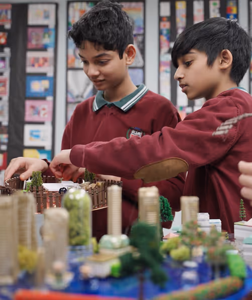 Two boys in maroon sweaters building a detailed city model with miniature buildings and trees.
