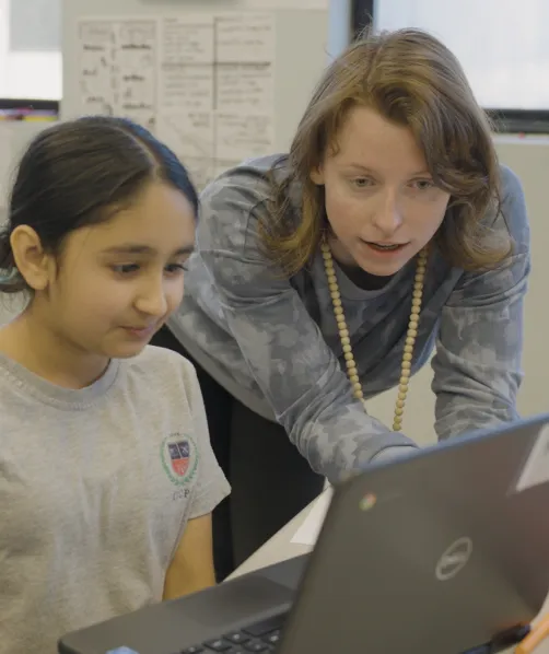 Teacher leaning over to assist a student working on a laptop in a classroom.