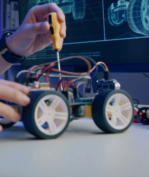 Person assembling a small robotic car with a screwdriver, with a computer screen displaying technical drawings in the background.