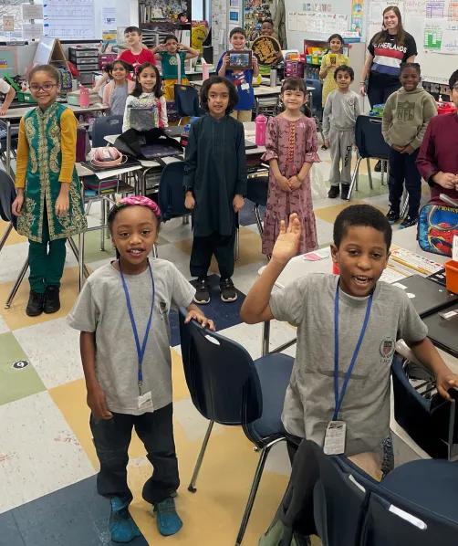 Diverse group of smiling elementary school children standing and sitting at desks in a colorful classroom.