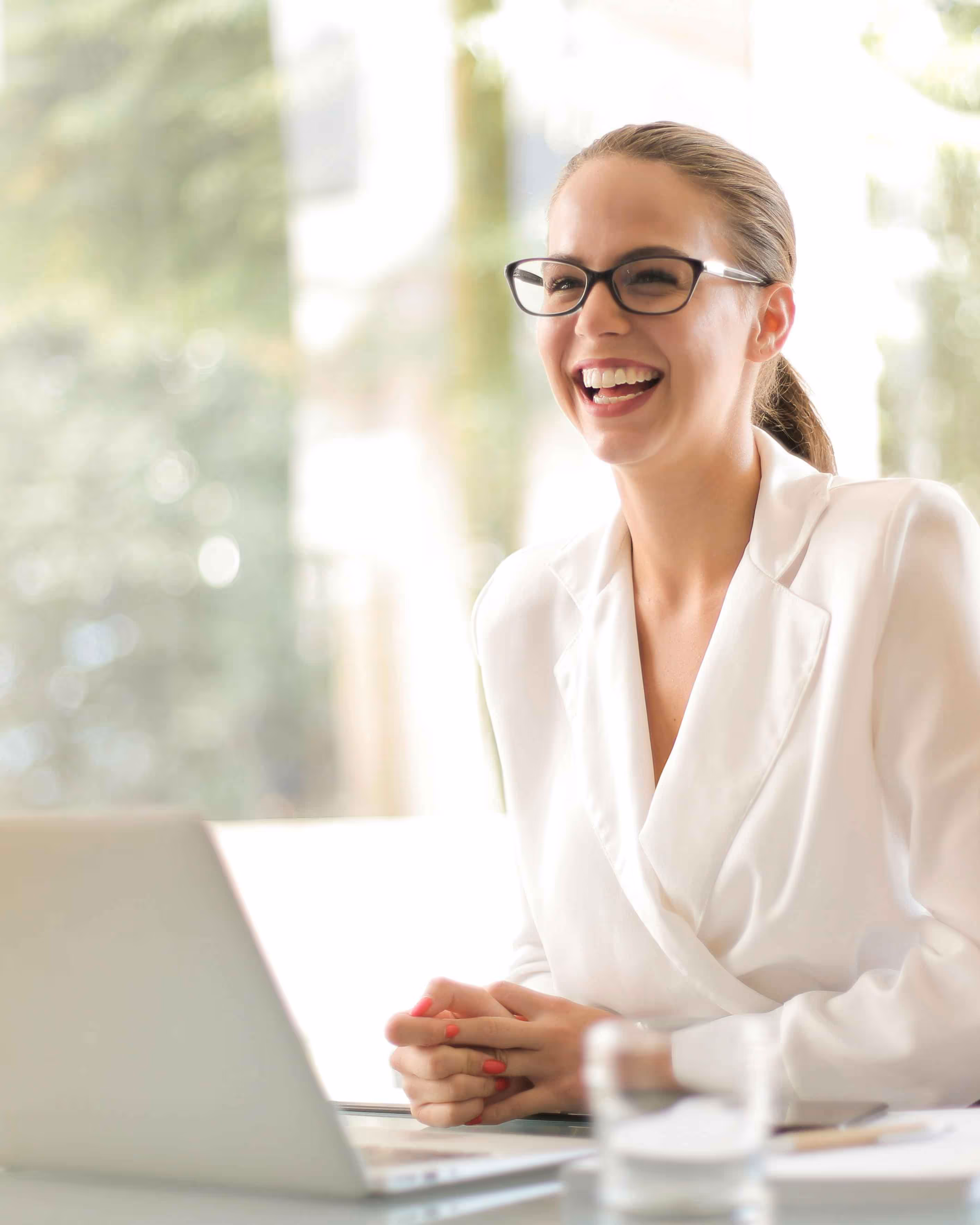 Smiling woman in white blazer wearing glasses sits at a desk with a laptop and a glass of water.