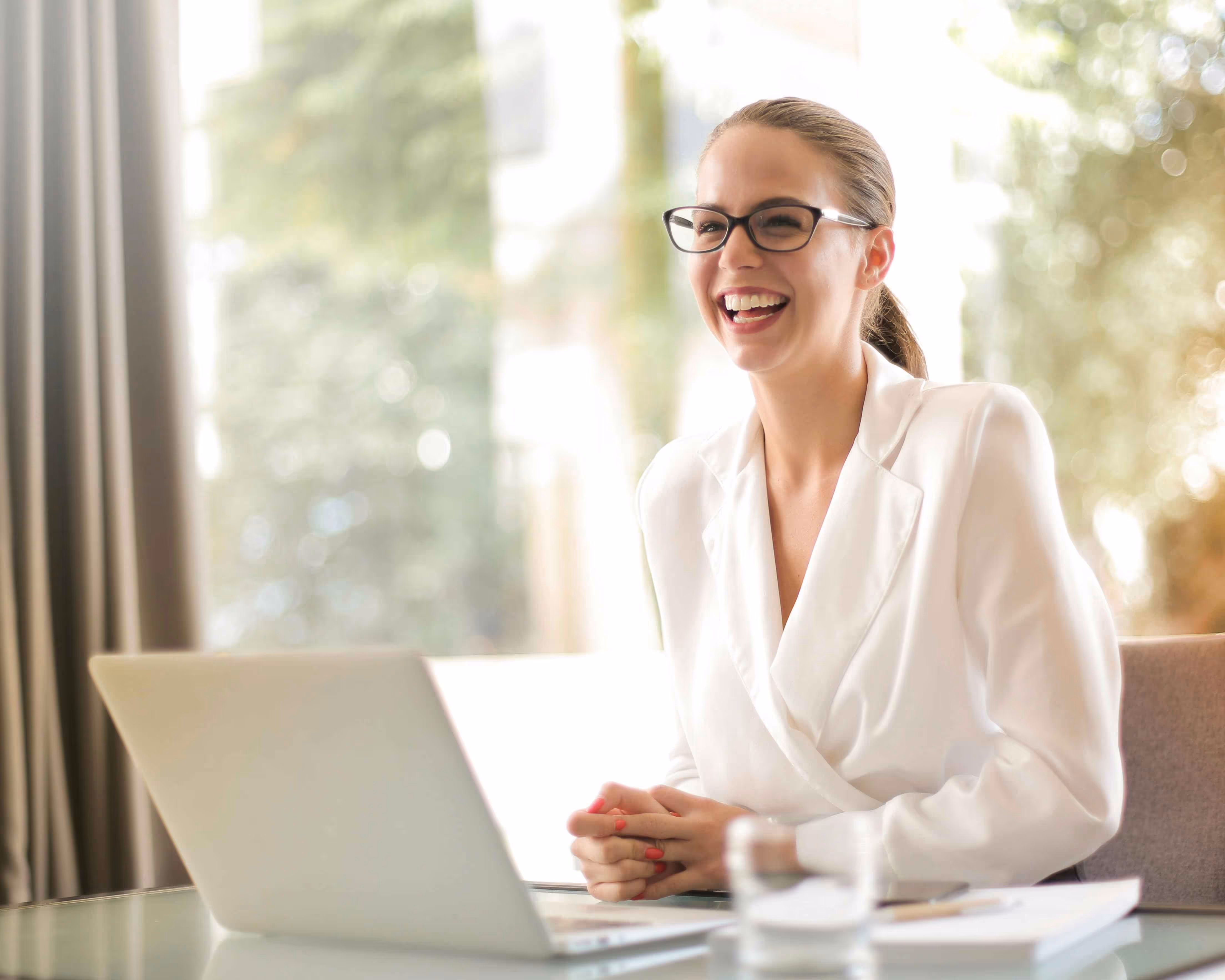 Smiling woman wearing glasses and a white blazer sitting at a desk with a laptop and documents.