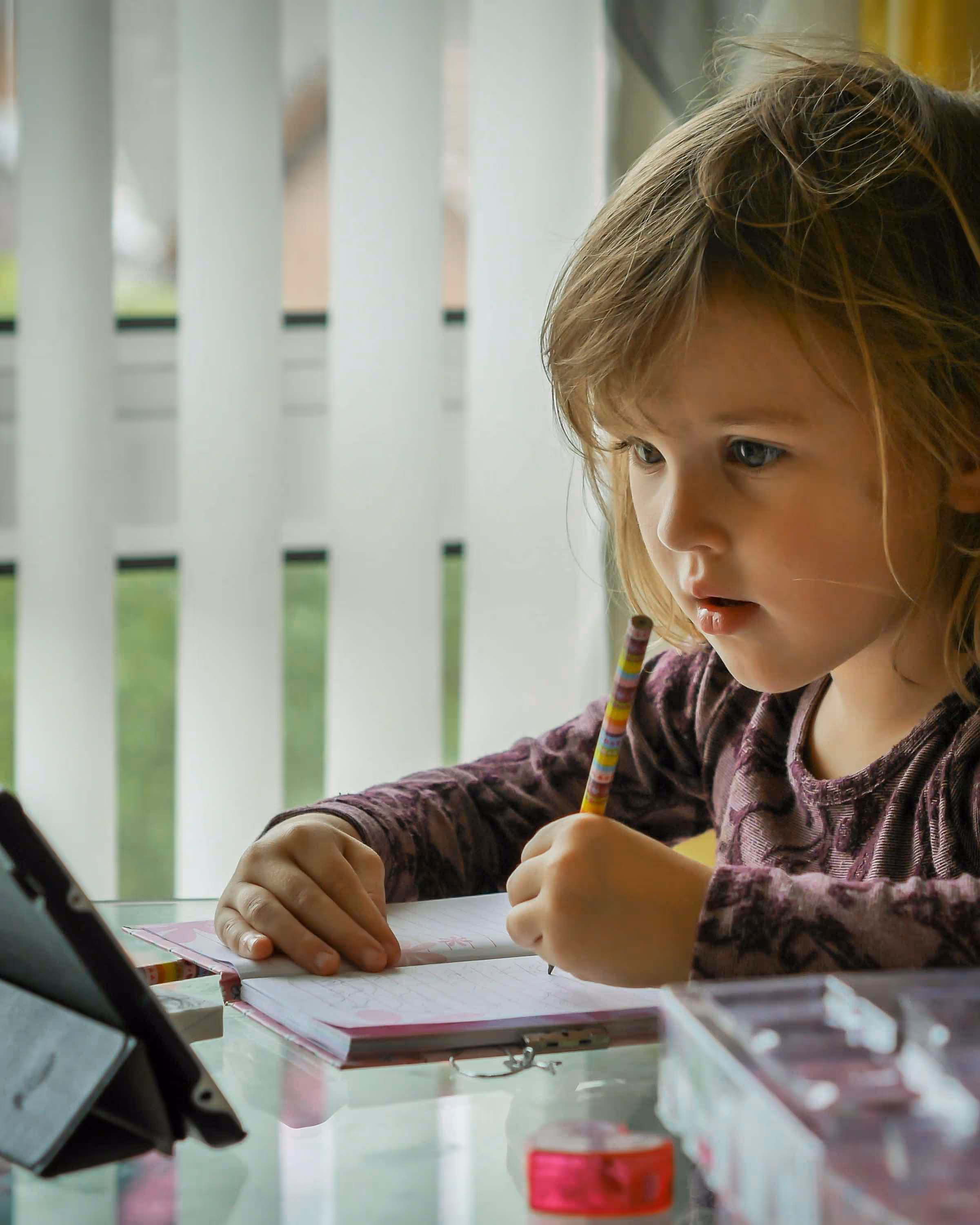 Young girl focused on writing in a notebook at a table with a tablet propped up nearby.