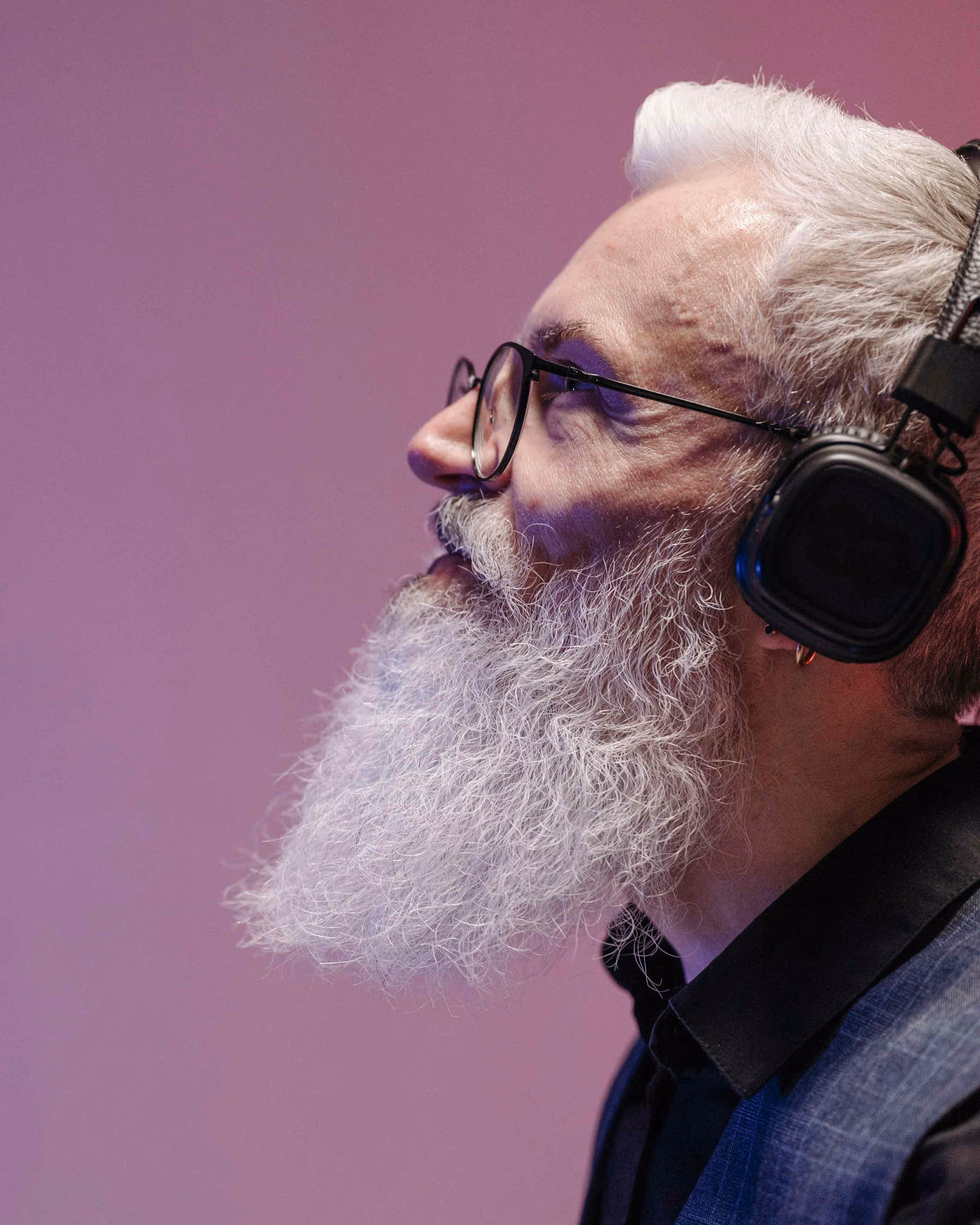Profile of an older man with white hair and a long white beard wearing glasses and black headphones against a purple background.