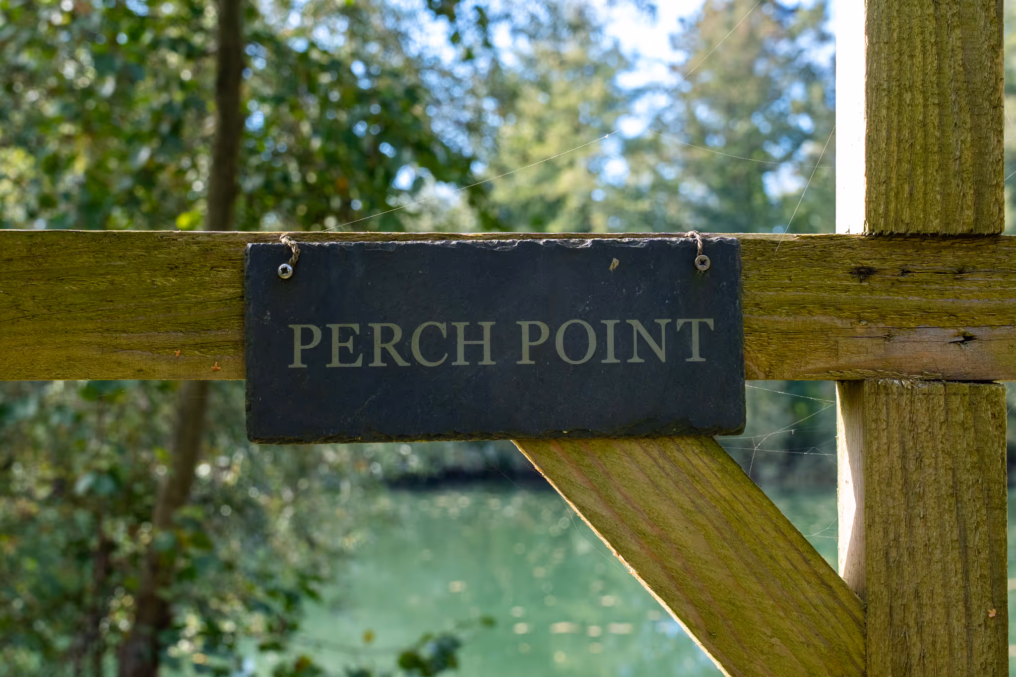 Panneau en ardoise portant l'inscription 'PERCH POINT' accroché à une barrière en bois avec un arrière-plan flou de verdure et d'eau.