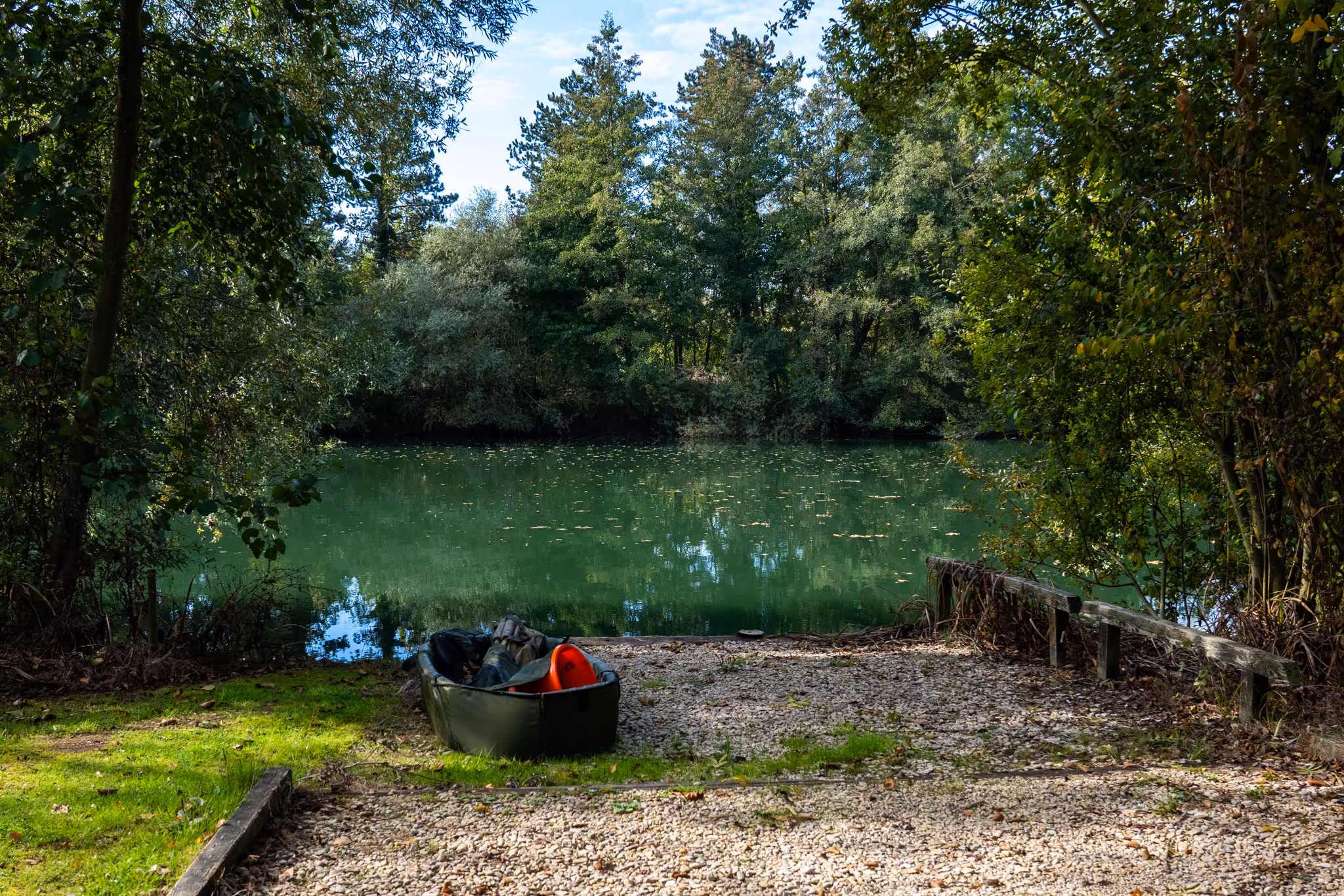 Canoë noir posé au bord d'un étang calme entouré d'arbres verts avec des feuilles flottant sur l'eau.