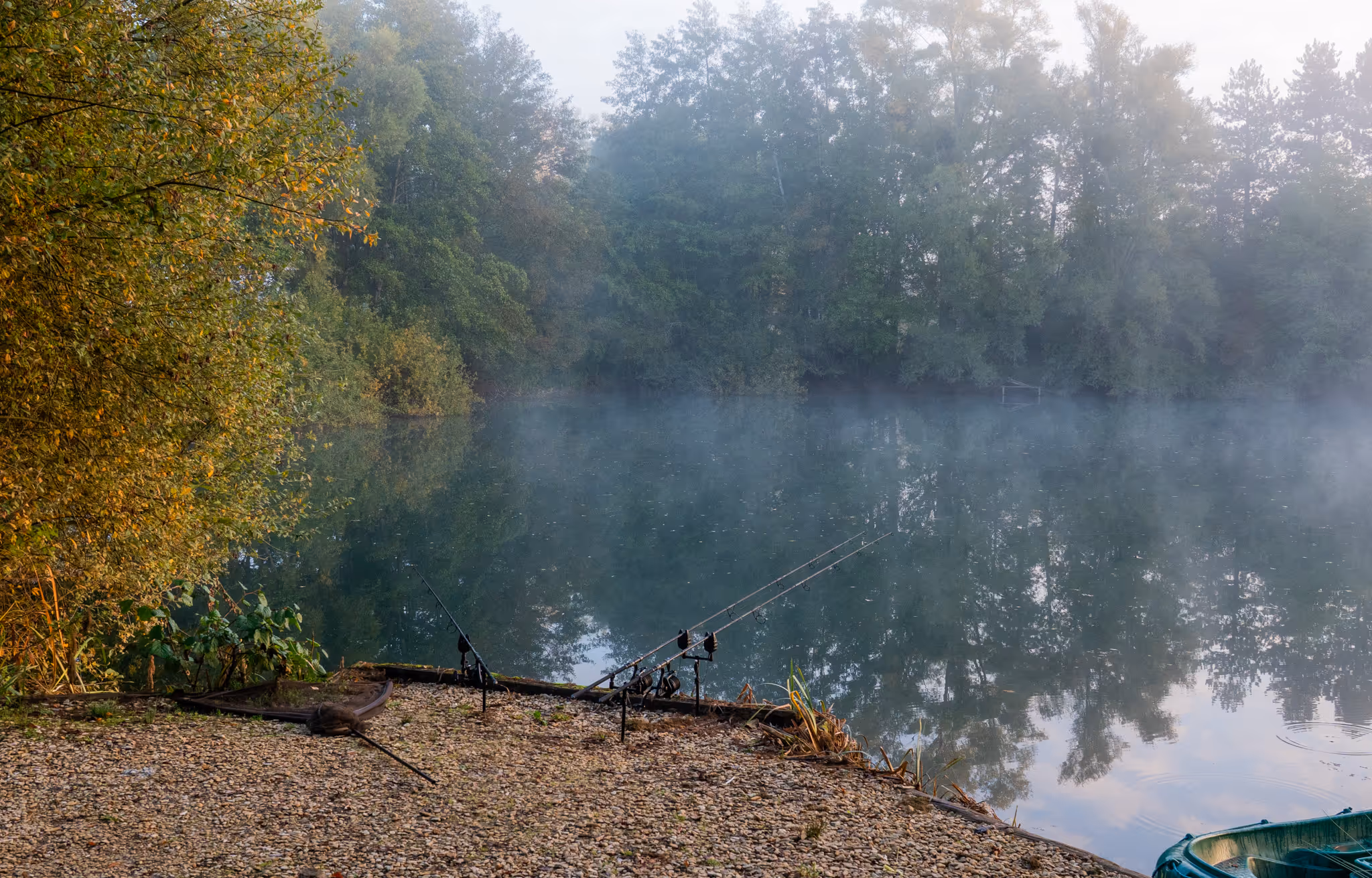 Deux cannes à pêche posées au bord d'un lac calme entouré d'arbres avec une légère brume sur l'eau et un petit bateau vert partiellement visible.