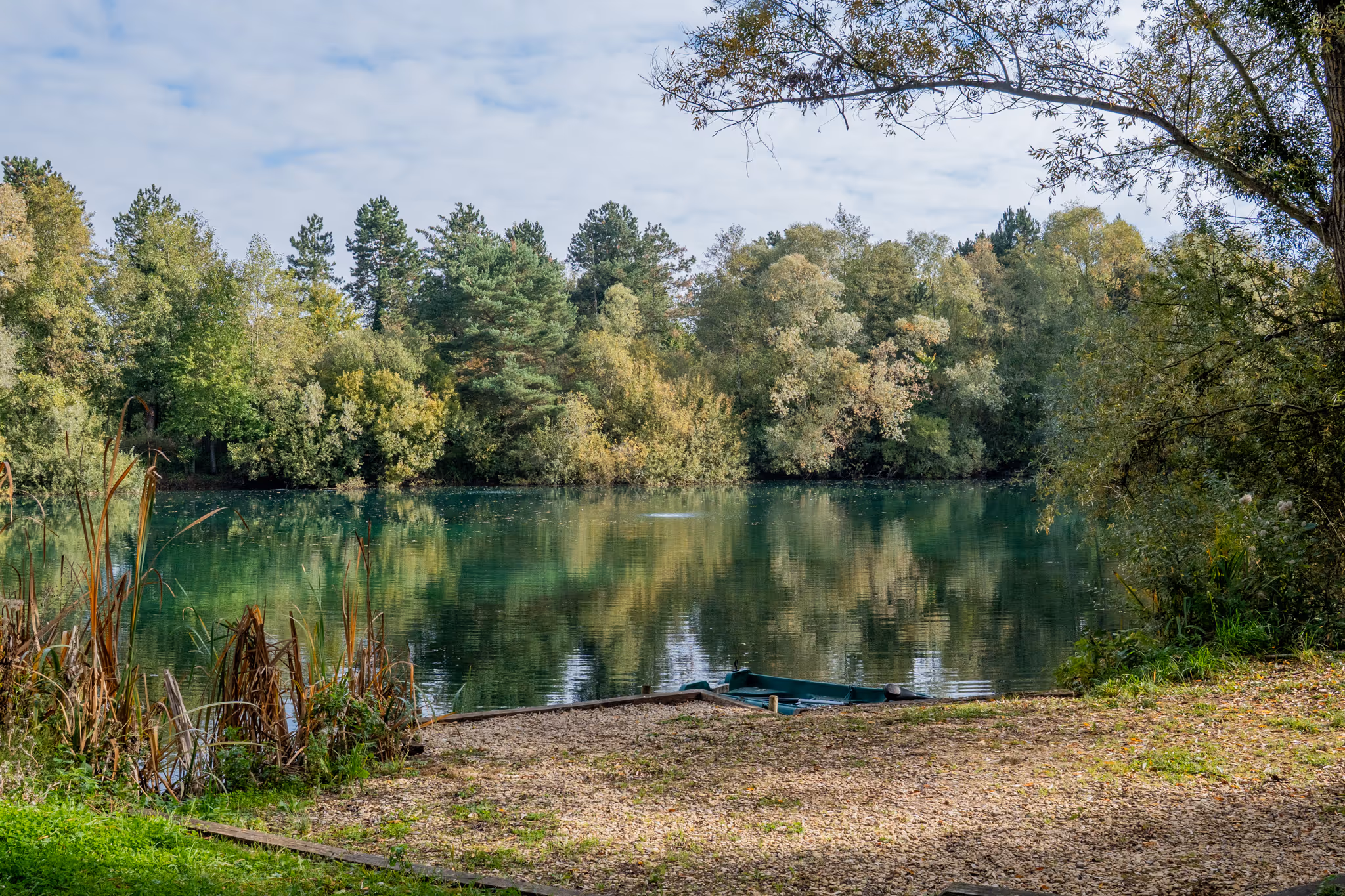 Petit quai en bois avec une barque verte sur un lac calme entouré d'arbres aux feuilles vertes et jaunes reflétées dans l'eau.