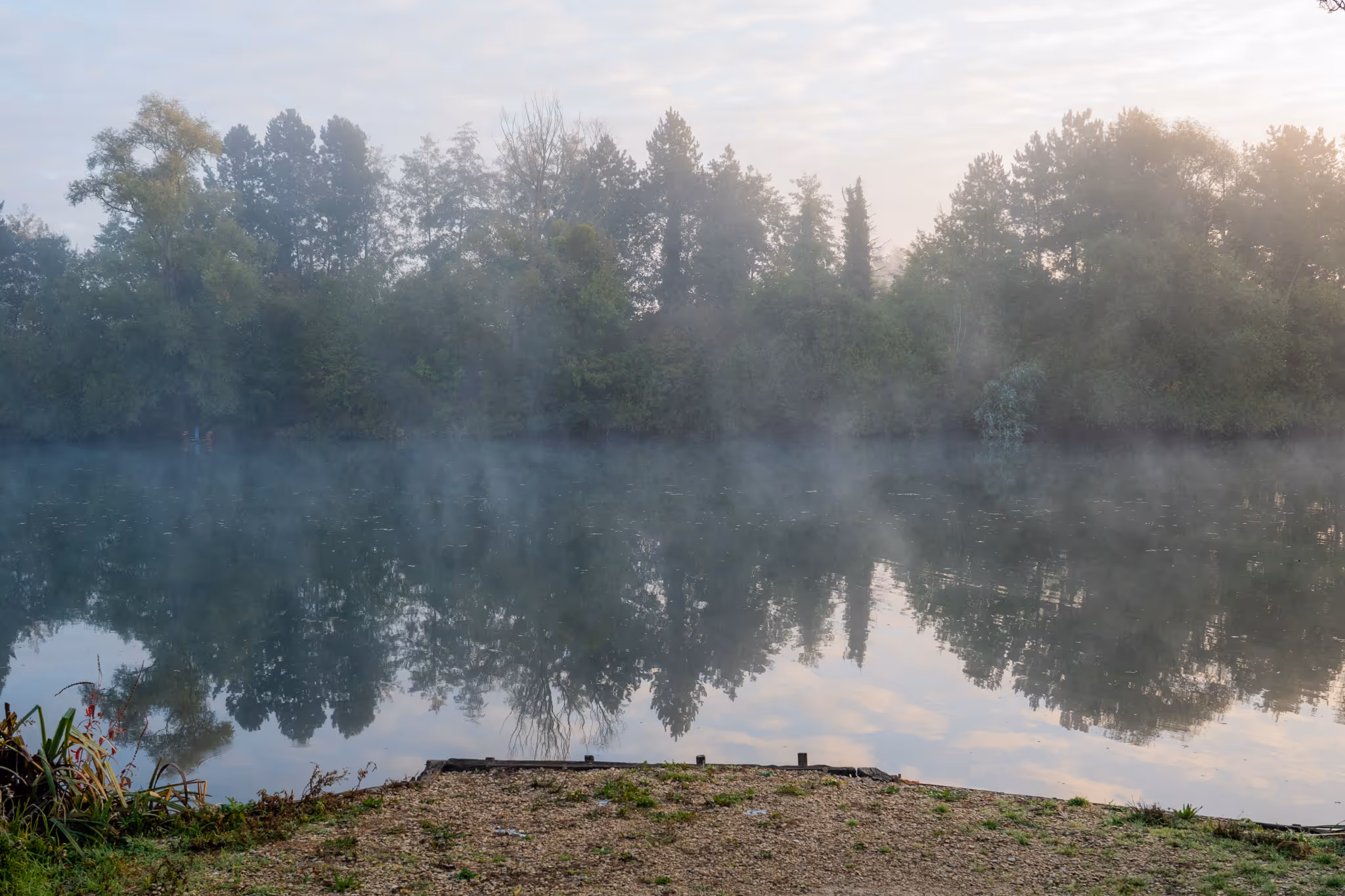 Un quai en gravier devant un lac brumeux reflétant des arbres verts sous un ciel nuageux au lever du soleil.