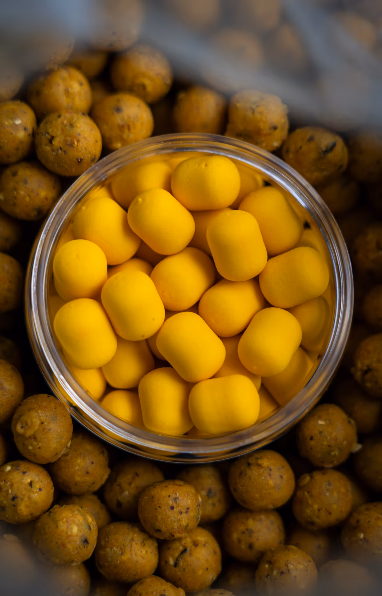 Un bol transparent rempli de petites boules jaunes allongées entouré de nombreuses boules rondes brunes.