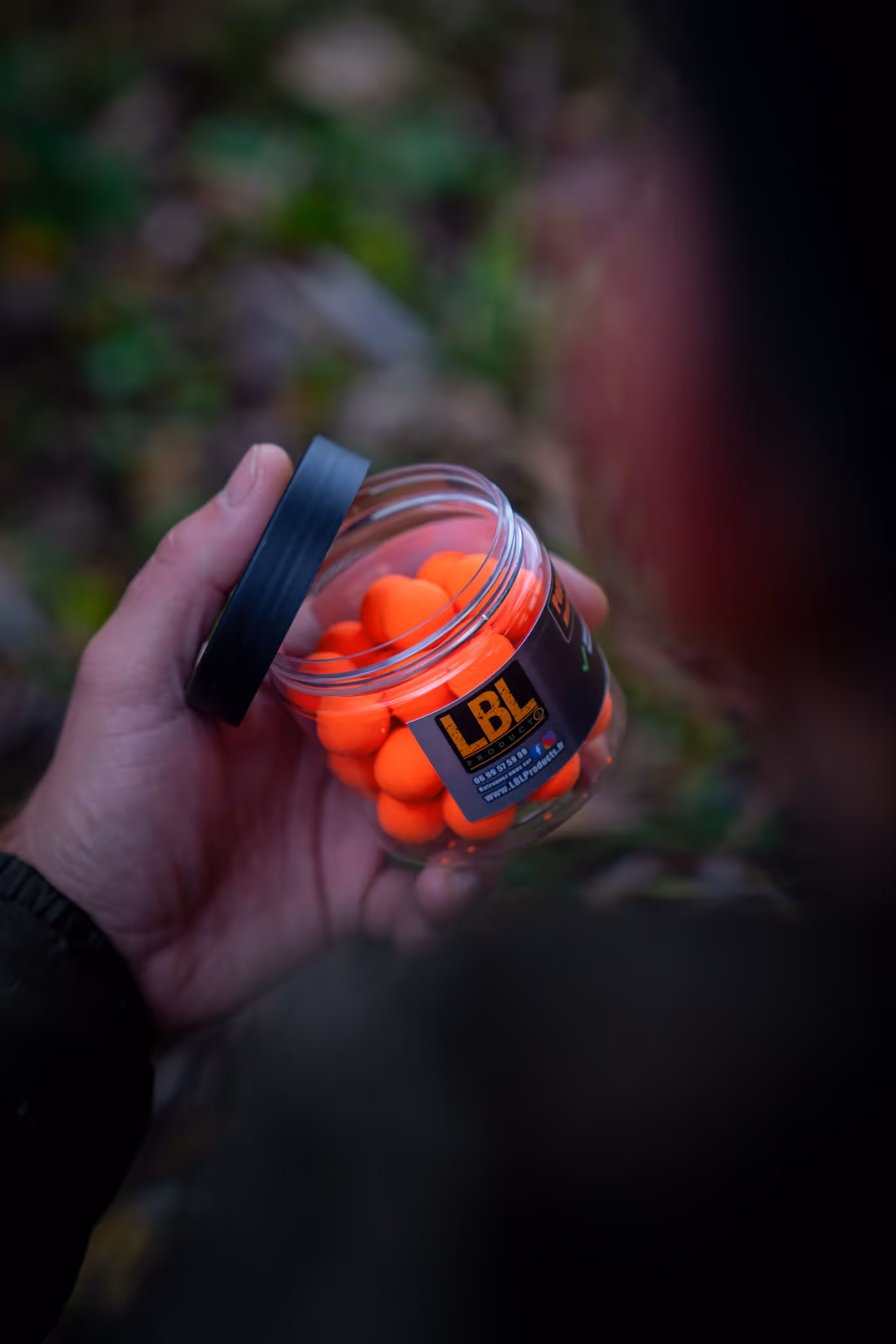 Main tenant un pot transparent contenant des boules orange avec un couvercle noir partiellement ouvert.