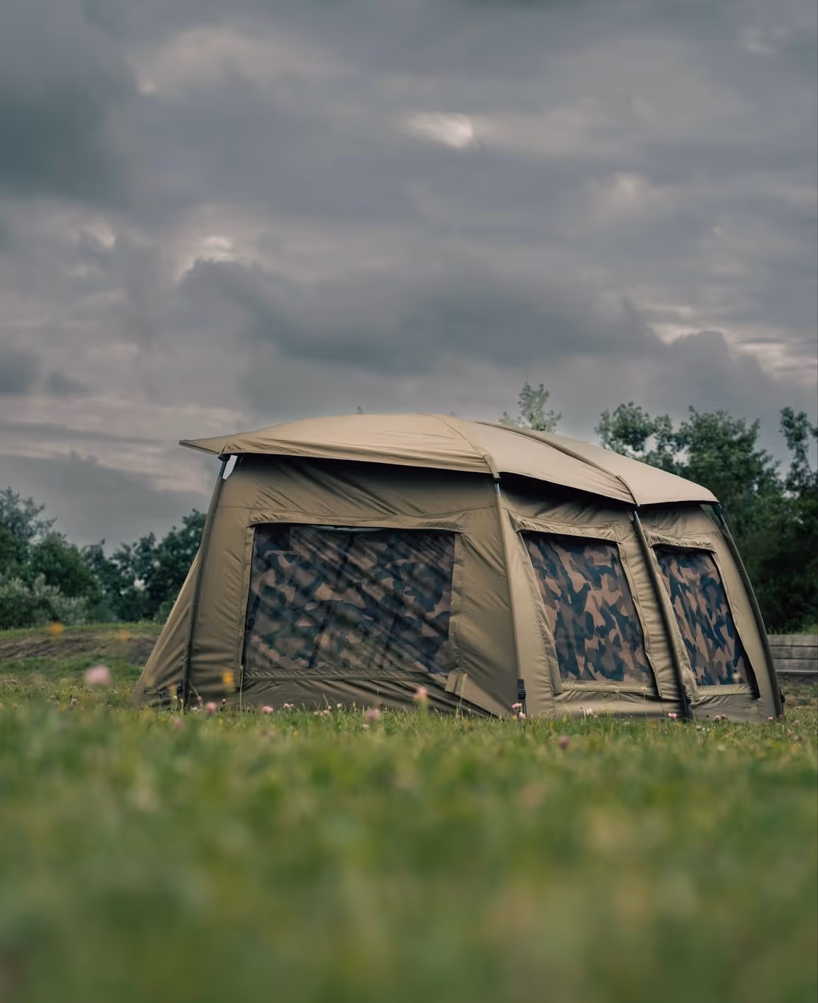 Tente kaki avec fenêtres camouflage installée sur une prairie sous un ciel nuageux.