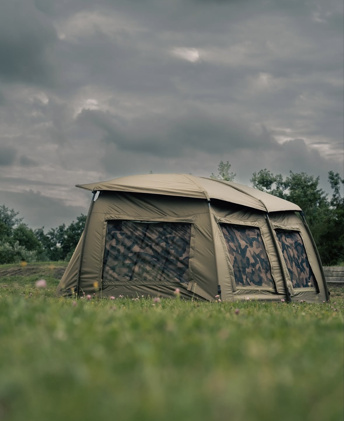Tente kaki avec fenêtres camouflage installée sur une prairie sous un ciel nuageux.