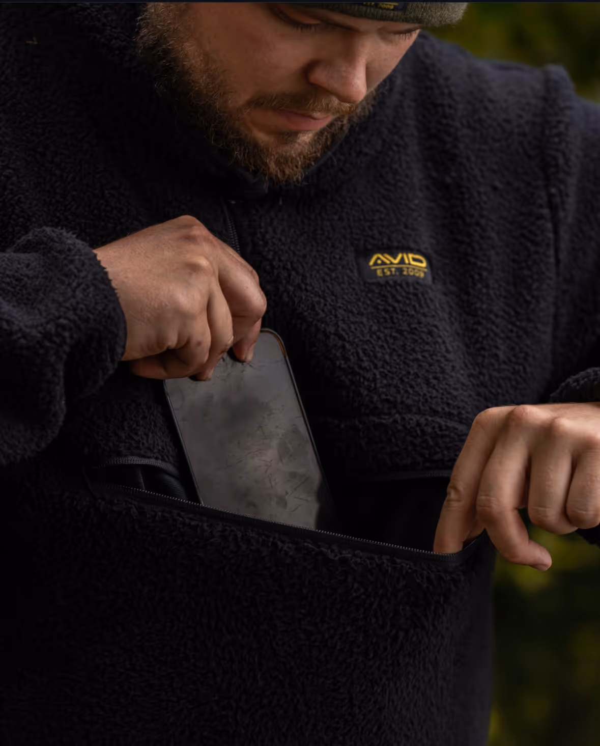 Un homme portant un pull en laine noire zippe une poche et y range une tablette avec un écran rayé.