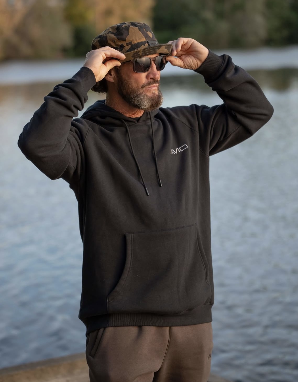 Homme barbu portant un sweat à capuche noir et un chapeau camouflage ajustant ses lunettes de soleil au bord de l'eau.