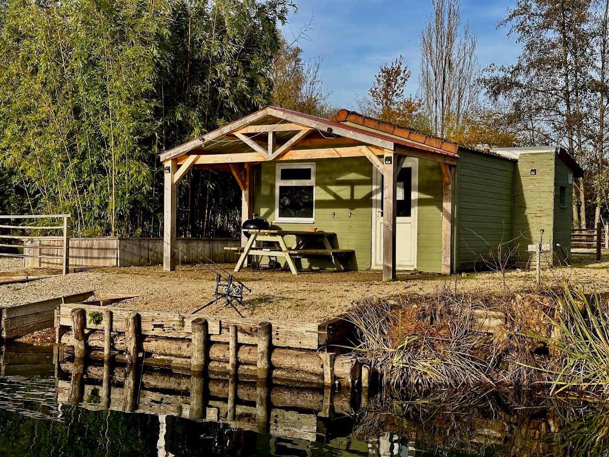 Cabane verte avec un porche en bois au bord d'un lac calme, équipée d'une table de pique-nique et d'un barbecue, avec des cannes à pêche installées devant.
