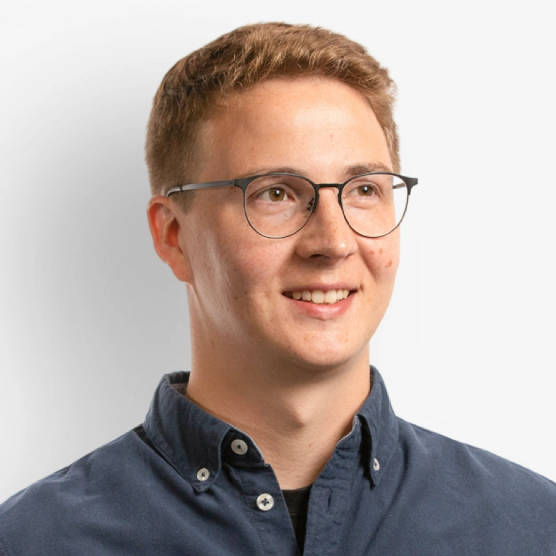 Young man with short light brown hair wearing round glasses and a dark blue button-up shirt smiling and looking slightly to the right against a white background.