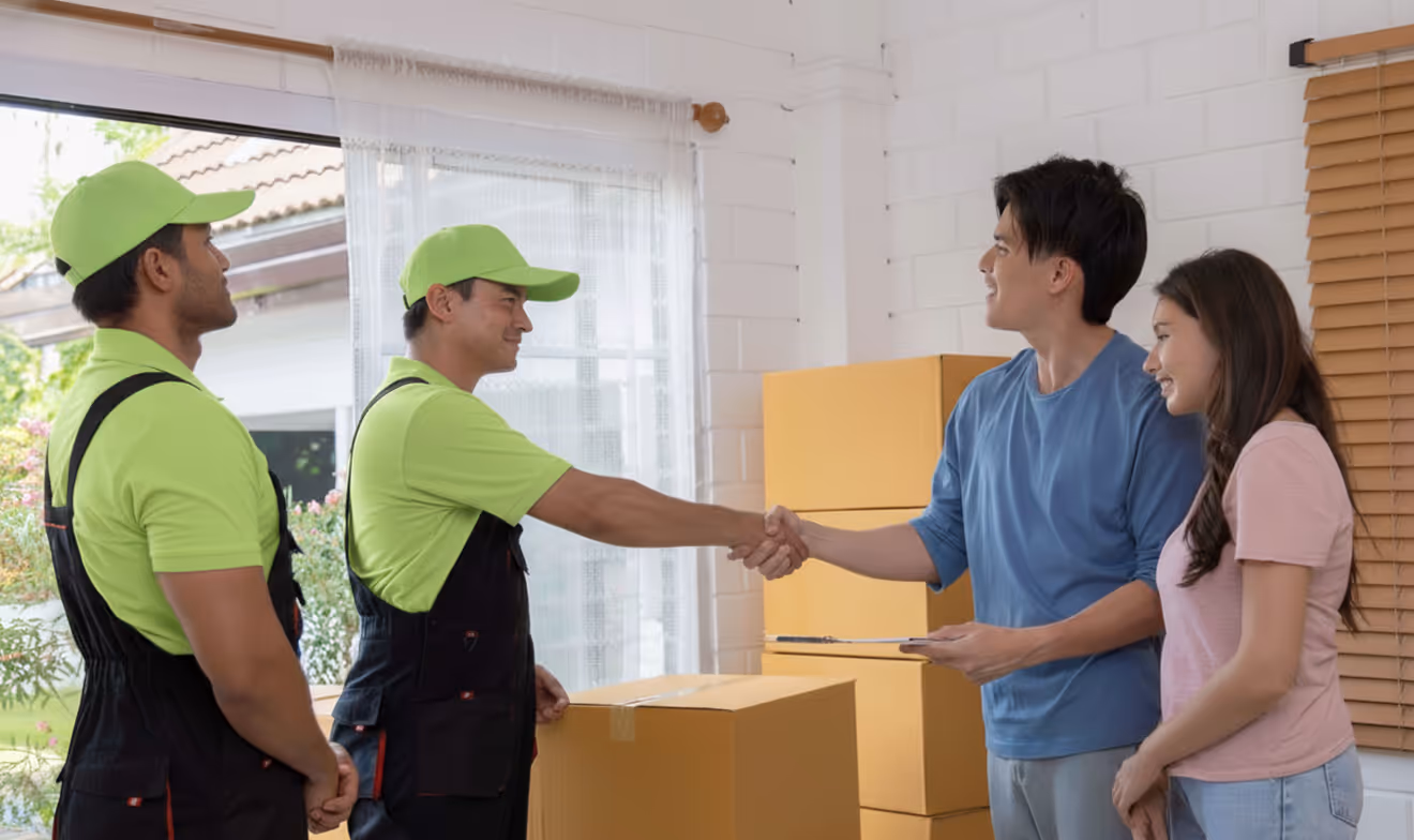 Delivery workers in green uniforms shaking hands with a man holding a clipboard as a woman stands beside him, with moving boxes stacked in the background.